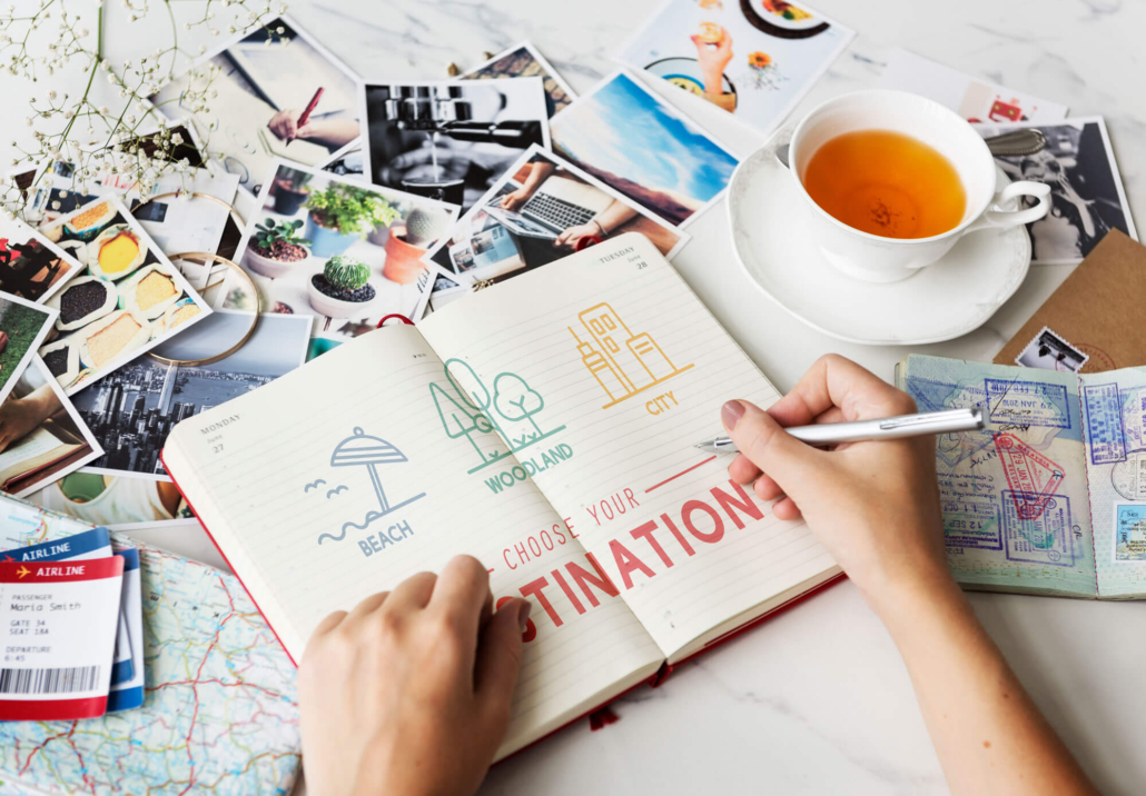 Auf einem Tisch voller Fotos liegt neben einer Teetasse ein Notizbuch in das gerade eine Person Reiseziele schreibt