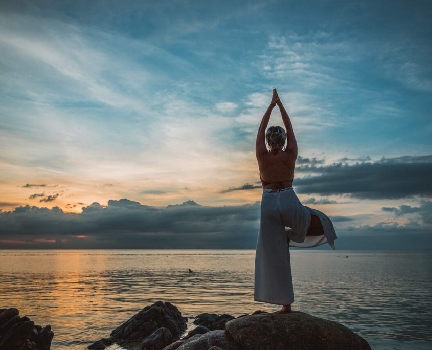 Eine Frau steht einbeinig an einem Ufer vor dem Sonnenuntergang und macht Yoga