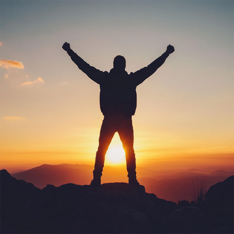 Eine Person steht auf einem Berg mit hochgerissenen Armen, zwischen seinen Beinen scheint der Sonnenuntergang hindurch